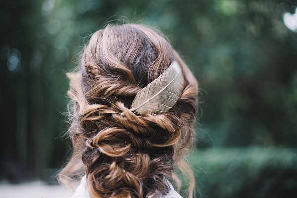 Close-up detail shot of a bride's updo hairstyle, photographed from behind. The hair is styled in a loosely braided and twisted updo with soft, wavy tendrils, accessorized with a single decorative feather hairpin in a muted gold or silver tone. The styling is consistent with a boho or bohemian wedding aesthetic. The subject is wearing a white garment visible at the bottom edge of the frame.