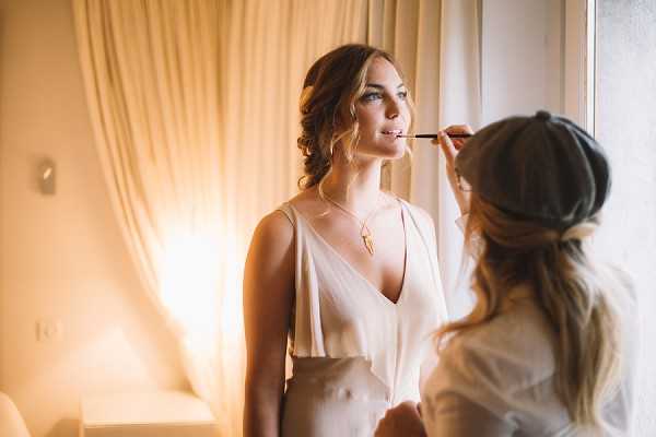 A bride is having her makeup applied by a makeup artist in an indoor getting-ready setting, likely a hotel room or bridal suite with warm ambient lighting and sheer cream curtains. The bride wears a sleeveless white V-neck top with a gold pendant necklace and has her blonde hair styled in a loose updo with soft waves. The makeup artist, wearing a grey flat cap, applies product with a brush while standing close to the bride near a window. The composition is a medium portrait shot with warm, golden-toned lighting creating a soft contrast against the natural light from the window.