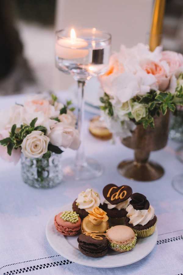 Close-up detail shot of a dessert display at a wedding reception, featuring a white plate arranged with an assortment of French macarons in blush pink, gold, and chocolate tones, mini cupcakes with white and dark chocolate frosting, and a chocolate heart decoration inscribed with 'I do' in gold lettering. The table is dressed in a white linen with delicate embroidered border detailing, and styled with blush pink and ivory rose arrangements in a silver mercury glass vase and a gold pedestal vase, accompanied by a tall glass floating candle holder with a lit candle and a gold taper candle in the background. The color palette combines blush, ivory, gold, and chocolate tones in a romantic classic style.