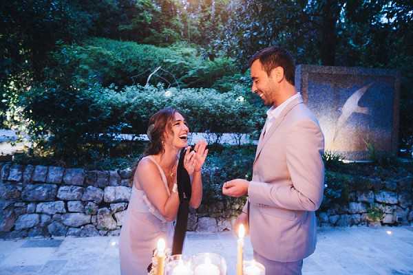 A couple shares a joyful, laughing moment outdoors at dusk in what appears to be a garden or courtyard setting, with a stone wall and decorative stone monument visible in the background. The woman wears a light silver-grey slip-style dress and a delicate necklace, while the man wears a light beige suit. Tall taper candles in glass holders are visible in the foreground, casting warm light against the blue evening atmosphere. The shot is a mid-range portrait capturing a candid, informal moment, likely during a proposal or intimate celebration.