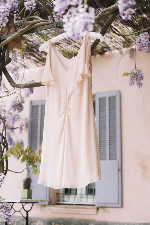 A blush pink bridesmaid or bride's short dress hangs on a white hanger from a wisteria-covered branch outdoors. The knee-length dress features a V-neckline, ruffle front detail, and a gathered cinch at the center bodice in a fluid, lightweight fabric. In the background, a pale peach rendered building with grey-blue shuttered windows is softly out of focus, with clusters of purple wisteria blooms framing the composition. This is a detail/close-up portrait-style shot styled as a getting-ready or flat-lay alternative image.