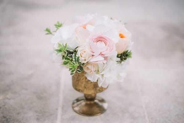 A close-up detail shot of a small floral arrangement displayed in a vintage brass footed vase or goblet. The arrangement includes blush pink ranunculus, cream roses, white peonies, and soft green foliage, styled in a compact rounded shape. The color palette is blush, cream, and white with gold accents from the vessel. The background is a softly blurred neutral stone or concrete surface, keeping full focus on the floral composition.
