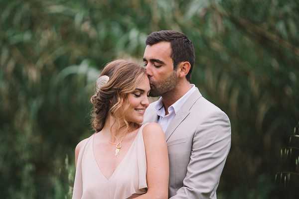 A couple portrait shot outdoors with the groom kissing the bride on the temple as she smiles with eyes closed. The bride wears a blush pink V-neck dress and has her hair styled in a loose updo adorned with a small gold shell or leaf hair accessory, paired with a delicate gold pendant necklace. The groom wears a light taupe/beige suit with an open-collar light blue shirt. The background is heavily blurred greenery, creating a soft bokeh effect. The image is a close-up portrait with warm, natural lighting.