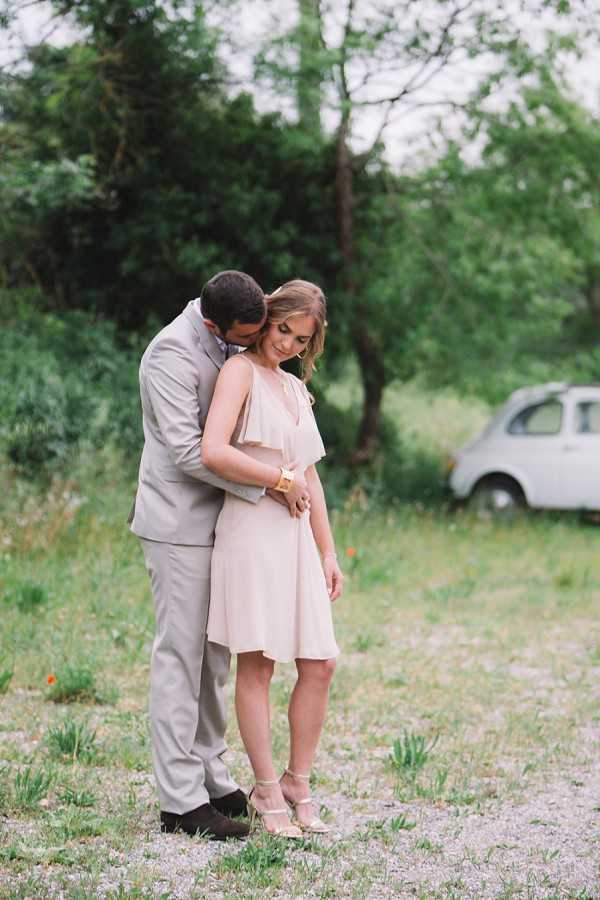 A couple portrait taken outdoors in a rural setting, with the man standing behind the woman and leaning in close to her. The man wears a light taupe/beige suit, and the woman wears a knee-length blush pink dress with a flutter neckline detail and strappy gold heeled sandals, accessorized with a gold bracelet. A vintage white compact car, resembling a Fiat 500, is visible in the soft-focus background among trees and low vegetation. The shot is a full-length portrait with a shallow depth of field, giving the background a blurred, painterly quality.