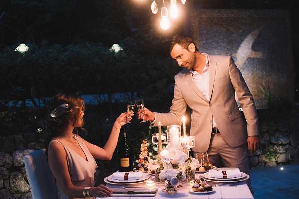 A couple toasts with champagne flutes at an intimate outdoor dinner table set for two during the evening. The groom wears a tan/beige suit with an open-collar white shirt, and the bride wears a blush or light pink sleeveless dress with her hair styled up and adorned with a decorative hair piece. The table is dressed with white linens, layered place settings with ribbon-tied napkins, pillar candles, a low floral centerpiece featuring white blooms, and a bottle of Champagne in the center. Warm Edison-style pendant bulbs hang overhead, and small dessert bites are visible on the table. The setting is an outdoor terrace or garden at night, with stone walls and garden lighting visible in the background, creating a dark, moody atmosphere. Medium portrait shot capturing both figures from approximately waist height.