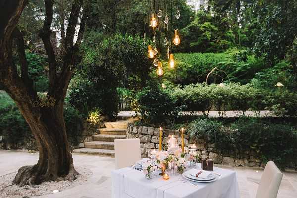 An outdoor sweetheart table setup in a garden setting, styled for a wedding reception with no people present. The small rectangular table is covered in a white linen and set for two, with white plates, glassware, and scattered low floral arrangements featuring blush and peach blooms with greenery. Tall amber taper candles and small votive candles provide warm light on the table surface. A cluster of Edison-style pendant bulbs and hanging crystal prisms suspended from the tree branches above creates a warm, ambient glow at dusk. Stone steps and terraced garden walls are visible in the background. The overall decor palette is white, gold, and blush with a modern-romantic styling. Wide shot composition.