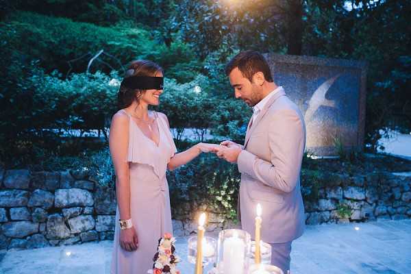 An outdoor evening scene showing a couple at what appears to be an intimate ring exchange or proposal moment, set in a garden or terrace area with stone wall backdrop and sculptural element. The woman wears a blush/taupe draped v-neck dress with a wrist cuff bracelet, while the man wears a light beige suit with no tie and an open-collar white shirt. Tall pillar candles and glass candleholders on a table in the foreground provide warm ambient lighting against the blue dusk sky. A small floral arrangement with deep red and blush flowers is partially visible at the table's edge. The shot is a medium portrait framing both figures from approximately the waist up.