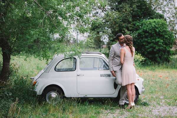 A couple shares a close, near-kissing moment leaning against a vintage white Fiat 500 with the word 'MAGAZZINO' printed on its door, positioned in an overgrown grassy outdoor setting. The man wears a light beige/tan suit and the woman wears a short blush pink dress with her hair styled in a loose braid. The shot is taken as a medium-wide portrait, capturing both figures full-length alongside the car. The overall styling is relaxed and vintage-inspired, pairing the retro vehicle with the couple's soft, warm-toned attire.