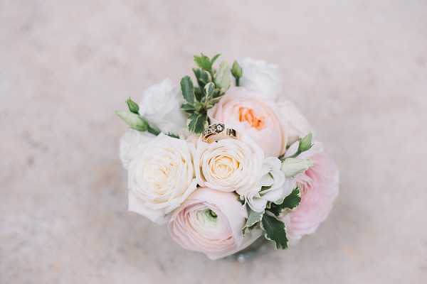 A close-up detail shot of a small bridal bouquet composed of blush pink garden roses, cream ranunculus, white lisianthus buds, and dusty miller foliage, placed on a pale marble surface. Two gold wedding rings — one a diamond engagement ring and one a plain band — are nestled among the blooms at the center of the arrangement. The bouquet palette is soft blush and cream with green accents. The image is sharply focused with a shallow depth of field and natural light.