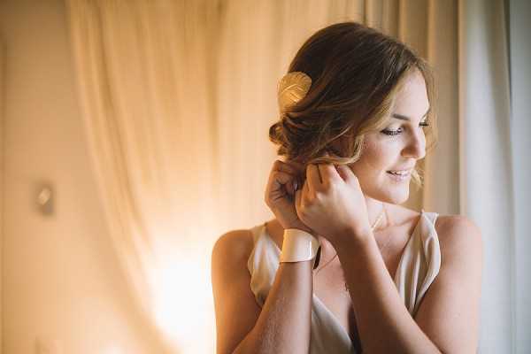 A bride is photographed getting ready, putting on an earring while smiling softly. She is indoors in front of sheer cream curtains that diffuse warm natural window light. She wears a sleeveless ivory or champagne slip-style top, a delicate pearl or crystal necklace, and a wide cream or ivory cuff bracelet, with her hair styled in a low updo accented by a gold leaf hair accessory. The shot is a close-up portrait with warm, soft-focus background tones, giving it an intimate, natural feel.