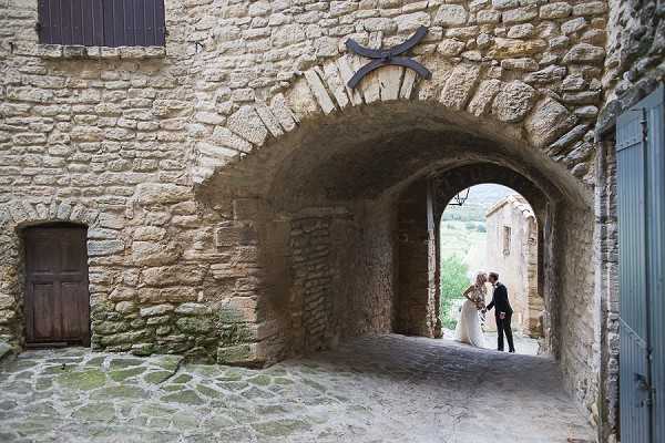 A couple shares a moment together beneath a stone archway passage in what appears to be a historic French village or chateau complex. The bride is wearing a white gown and holding a bouquet, while the groom is dressed in a dark suit. The wide shot frames them at the far end of a vaulted stone tunnel, with a second archway visible beyond them and greenery glimpsed in the background. The composition uses the deep archway architecture as a natural frame, placing the couple as small figures within the grand scale of the medieval stonework. Potential venue feature image.