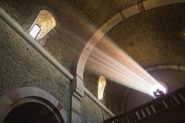 A wide-angle architectural interior shot taken from a low angle looking up at the vaulted stone ceiling of a Romanesque chapel or church. Strong rays of natural light stream dramatically through a large arched window at the upper right, illuminating the rough-hewn stone barrel vault and casting visible light beams across the space. Two small figures — appearing to be the couple — are silhouetted in front of the bright window, creating a stark contrast against the intense backlight. The stone walls feature arched niches and additional narrow windows, with pale trim detailing along the arches. The composition emphasizes the architectural grandeur of the space over the human subjects. Potential venue feature image.