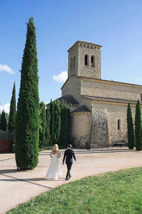 A bride and groom walk together along a gravel path toward a Romanesque stone chapel with a square bell tower, photographed from behind in a wide shot. The bride wears a fitted, low-back white lace gown with a short train, and the groom is dressed in a dark suit; he carries a dark bouquet. Tall, narrow Italian cypress trees line the path on either side of the chapel, framing the building. The setting is outdoor and rural, consistent with a South of France or Provence landscape, with a classic, understated styling approach. Potential venue feature image.