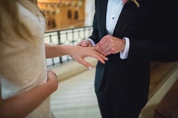 Close-up detail shot of a ring exchange moment during an indoor wedding ceremony. The groom, dressed in a black tuxedo with a white dress shirt and boutonniere, is placing a wedding band onto the bride's finger; the bride is wearing a lace or embroidered white dress with cap sleeves. The setting appears to be an ornate interior space with a decorative iron railing and warm stone or marble flooring visible in the background.