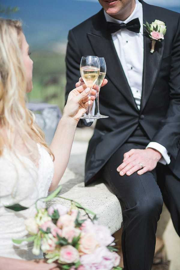 The bride and groom share a champagne toast while seated outdoors on a stone ledge or bench. The groom wears a black tuxedo with a black bow tie and a blush pink rose boutonniere, while the bride is in a white lace gown with wavy blonde hair. Their champagne flutes clink together in the foreground, and the bride holds a loose bouquet of blush pink and cream roses with greenery resting in her lap. The shot is a close-up portrait framed from mid-torso, emphasizing the toast and floral details.