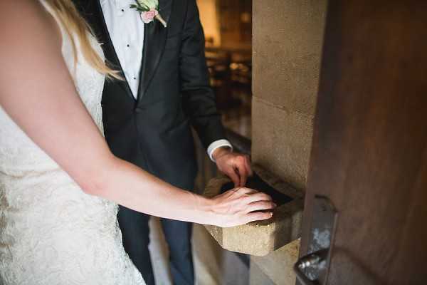 Close-up detail shot of a bride and groom's hands resting on a stone holy water font at the entrance of a chapel or church, likely following or preceding a religious ceremony. The bride, visible from the shoulders down, wears a lace white dress, while the groom wears a dark navy suit with a white dress shirt and a pink floral boutonnière. Wooden pews are softly visible in the background, confirming an indoor religious setting. The composition focuses on the hands and the carved stone basin, emphasizing a quiet ceremonial moment.