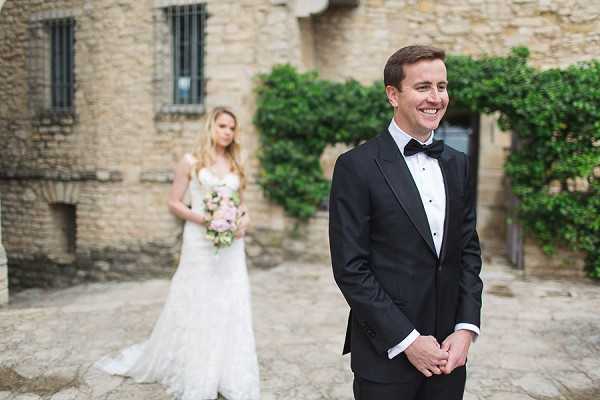 A first-look moment captured in the courtyard of a stone chateau or historic building, with the groom standing in the foreground smiling broadly with his hands clasped, dressed in a black tuxedo with a black bow tie and white shirt. The bride stands softly out of focus in the background wearing a fitted lace gown with a sweetheart neckline and a short train, holding a blush pink and white bouquet with greenery. The medium-wide portrait shot uses shallow depth of field to keep focus on the groom's expression while the bride remains visible behind him. The setting features aged stone architecture with climbing green ivy, giving the scene a classic French château feel.
