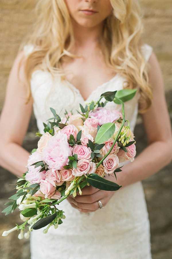 A close-up portrait of a bride holding her bridal bouquet, with the frame cropped just above her lips to just below her hips. She wears a fitted ivory lace dress with a sweetheart neckline and cap sleeves, and has long wavy blonde hair. The bouquet is composed of blush pink peonies, light pink spray roses, peach garden roses, white freesia, and loose greenery including eucalyptus and broad-leaf stems that trail downward in a loosely arranged, slightly cascading style. A diamond engagement ring is visible on her left hand. The background is a warm-toned stone wall, softly blurred. The overall styling is classic and romantic with a soft blush and ivory palette.