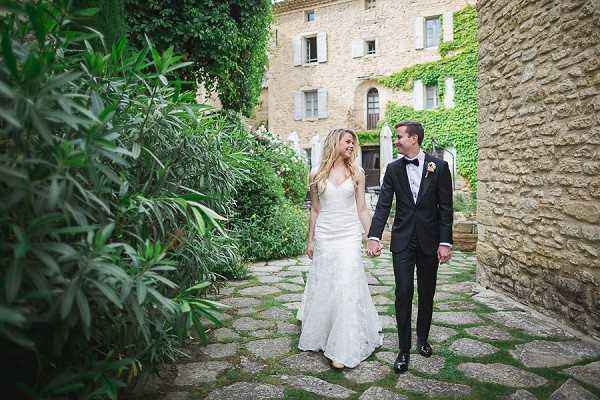 A bride and groom walk hand-in-hand along a cobblestone courtyard path between stone walls of a Provençal-style chateau covered in ivy. The bride wears a fitted, sleeveless lace gown in ivory with a V-neckline, while the groom is dressed in a black tuxedo with a bow tie and a small floral boutonniere. The shot is a medium-wide portrait capturing the couple from head to toe as they look at each other and smile. Potential venue feature image.
