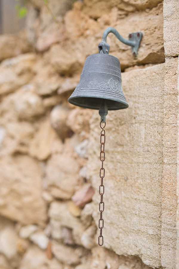 Close-up detail shot of a vintage cast-metal bell with a verdigris patina, mounted on a carved stone wall via a curved iron bracket, with a rusted link chain hanging below it. The bell features decorative floral engraving around its base. The background shows a rustic dry-stone wall softly blurred through shallow depth of field. No people are present; this appears to be an architectural or venue detail shot at an outdoor French property with rustic character. Potential venue feature image.