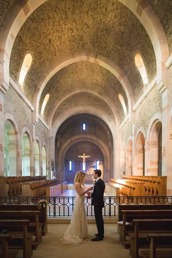A bride and groom exchange rings during their wedding ceremony inside a Romanesque stone chapel with barrel-vaulted ceilings, rounded arches, and a crucifix visible at the far end of the nave. The couple stands at a wrought-iron railing near the altar area, facing each other, with rows of wooden pews visible on either side. The bride wears a fitted lace gown with a short train, and the groom is dressed in a dark suit with a white boutonniere. The interior is warmly lit, highlighting the textured stonework of the arched ceiling, and a small floral arrangement in warm tones is visible near the railing. Wide-shot composition captures the full length of the chapel nave, emphasizing the architectural depth and scale of the space.