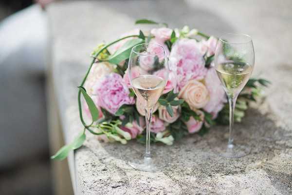 A close-up detail shot of two champagne flutes filled with sparkling wine placed on a stone surface in front of a bridal bouquet. The bouquet is composed of blush pink peonies, light pink spray roses, and trailing greenery including eucalyptus. The styling palette is soft blush and pale pink with green accents, consistent with a classic romantic aesthetic. The background is softly blurred, keeping focus on the glasses and florals.