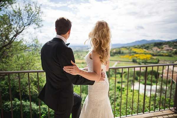 A couple stands together on an outdoor terrace or balcony with a wrought-iron railing, looking out over a sweeping rural landscape with rolling hills and mountains in the distance. The groom wears a dark charcoal suit with a bow tie, while the bride wears a fitted lace gown with a low back, her long blonde wavy hair loose. She has her hand placed on his lower back as they stand side by side. The shot is taken from behind in a wide portrait composition, emphasizing the panoramic countryside view below.