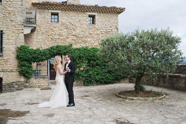 A couple shares a private moment in the cobblestone courtyard of a rustic stone-built chateau or mas, shot as a wide portrait. The bride wears a fitted, low-back ivory lace gown with a small train, and the groom is dressed in a classic black tuxedo with a white dress shirt and black bow tie. They are facing each other and appear to be in quiet conversation, standing in front of the building's ivy-framed entrance. The setting has a classic Provençal feel with aged stone architecture, a wrought-iron balcony, and a mature olive tree in a circular stone planter to the right. Potential venue feature image.