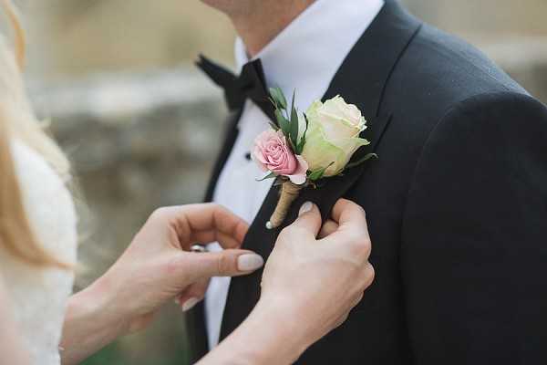 A close-up detail shot of a bride's hands pinning a boutonniere onto a groom's lapel. The groom is wearing a black tuxedo jacket with a white dress shirt and black bow tie. The boutonniere features a cream rose and a smaller dusty pink rose, accented with small green leaves and wrapped with twine at the stem. The bride's left hand is partially visible, showing a ring. The background is softly blurred with neutral stone tones. The overall styling is classic and formal.