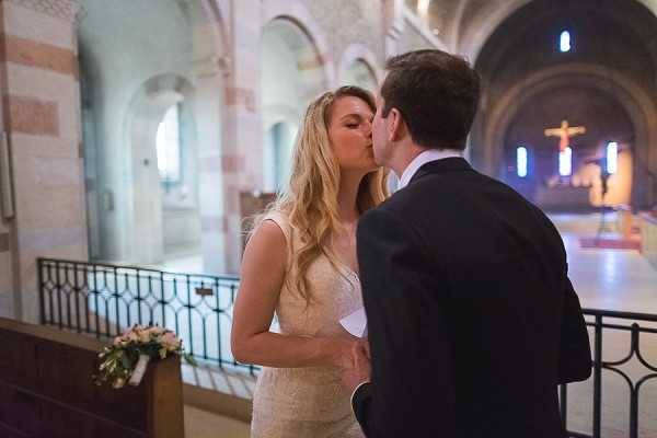 A bride and groom share their first kiss during an indoor church ceremony. The bride has long blonde hair and wears a fitted ivory lace gown, holding a small bouquet with pale pink flowers, while the groom is in a dark navy suit. The setting is a Romanesque-style stone church with rounded arches, striped stonework in beige and pink tones, and a crucifix illuminated in blue and amber light visible in the background. The shot is a medium portrait taken from the side, with the church interior and empty pews slightly blurred in the background.