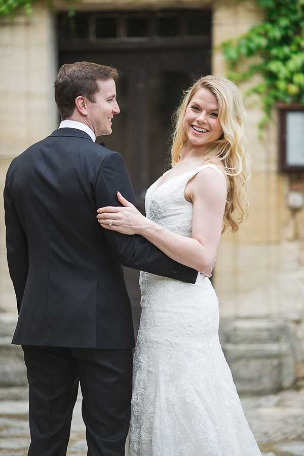 A couple portrait taken outdoors in front of a stone building with a large wooden door and ivy-covered walls. The bride, wearing a fitted white lace gown with a V-neckline and her blonde hair worn loosely in waves, smiles broadly toward the camera while placing her hand on the groom's back. The groom, dressed in a black suit with a white dress shirt, is turned slightly away from the camera and faces the bride. The shot is a medium portrait composition capturing both figures from approximately the knees up.