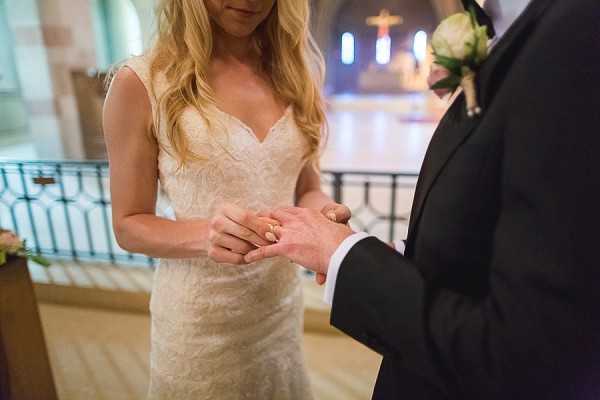 A close-up portrait shot of the ring exchange moment during an indoor religious ceremony, showing the bride placing a gold ring on the groom's finger. The bride has long blonde hair and wears a sleeveless, V-neck fitted lace gown in ivory, while the groom wears a dark navy or black suit with a white and blush boutonniere. The ceremony takes place inside a church, with a warmly lit altar area visible in the background featuring a crucifix and blue-toned stained glass or lighting. The composition is a medium close-up focused on the couple's hands and torsos, with the church interior softly blurred behind them.