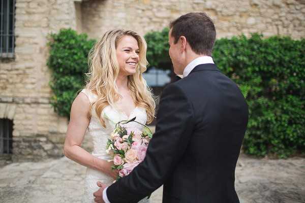 A couple shares a candid moment outdoors in front of a stone building, likely during a first look or post-ceremony portrait session. The bride, with long wavy blonde hair, wears a fitted ivory lace dress with a sweetheart neckline and holds a bouquet of soft pink roses and peonies with greenery. The groom, seen from behind, is dressed in a black suit with a white dress shirt. The setting appears to be a French stone courtyard or chateau exterior, with trimmed hedges visible in the background. This is a close-up portrait shot capturing the bride's natural smile as she faces the groom.