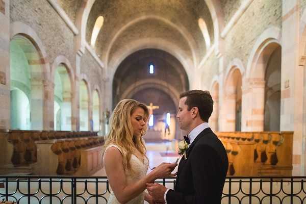 A bride and groom exchange rings during their ceremony inside a Romanesque stone church, characterized by rounded arches, barrel-vaulted ceilings, and rows of dark wooden pews visible in the background. A crucifix is positioned at the far end of the nave, illuminated by warm light filtering through stained glass. The bride wears a fitted lace gown with thin straps and has long blonde wavy hair, while the groom is dressed in a black tuxedo with a white boutonniere. The shot is a medium close-up portrait framing both figures from the waist up.