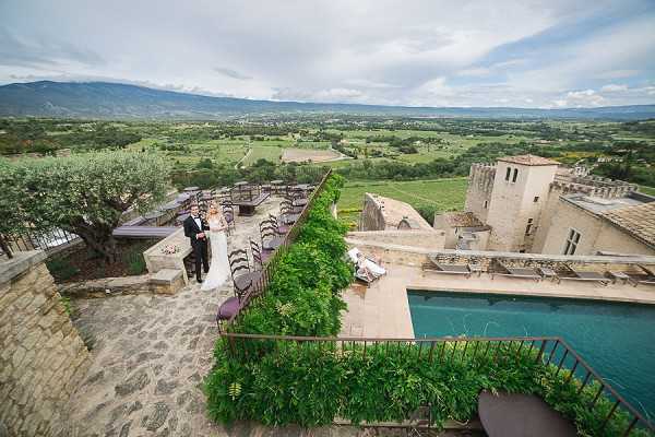 An aerial wide shot of a bride and groom posing on a stone terrace of a historic chateau, with a sweeping valley landscape visible in the background. The venue features a rectangular pool surrounded by greenery, a separate terrace area with rows of dark chairs set for an outdoor ceremony or dining, and medieval stone tower architecture. The bride wears a white gown and the groom a black tuxedo; they stand near an olive tree on the upper terrace level. The decor is minimal in this shot, with the focus on the architectural layout of the property and its elevated hillside position. Potential venue feature image.