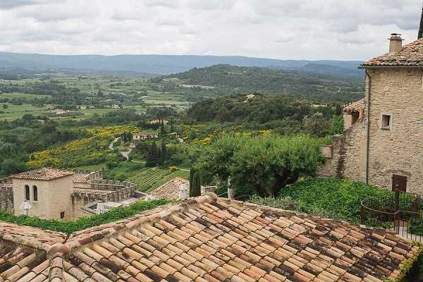 Wide-angle landscape shot taken from an elevated position at what appears to be a historic stone property in the south of France, showing terracotta-tiled rooftops in the foreground and a sweeping valley view in the background. The stone buildings visible include a structure with arched architectural details and what appears to be a classical white stone statue on a terrace or balustrade. No people or wedding party are visible in this image. Potential venue feature image.