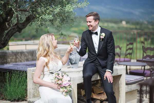 A bride and groom share a champagne toast while seated on a stone outdoor terrace, likely during a post-ceremony portrait session or cocktail hour. The bride wears a white lace cap-sleeve gown and holds a loose bouquet of pink and blush blooms including what appear to be ranunculus and garden roses with greenery; the groom wears a black tuxedo with bow tie and a pink and white boutonniere. The setting is an outdoor terrace with stone seating and a low stone wall, with a vineyard landscape and rolling hills visible in the background. The image is a mid-range portrait shot with natural daylight, reflecting a classic French countryside aesthetic.