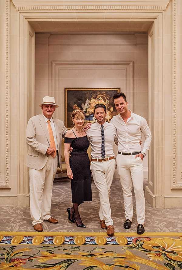 Four people pose together in a formal interior hallway or reception area, likely during a wedding cocktail hour or reception. The setting features white molded paneling, an ornate patterned carpet with gold and floral motifs, and a large oil painting visible in the arched alcove behind the group. The group consists of an older man in a cream suit with an orange tie and white panama hat, a woman in a black off-the-shoulder midi dress with black heels, and two younger men — one in cream trousers, a white dress shirt and polka-dot tie with a tan belt, and the other in an all-white shirt and trousers with a white belt. The overall style of the event appears classic and formal. This is a mid-range portrait shot capturing the full figures of all four subjects.