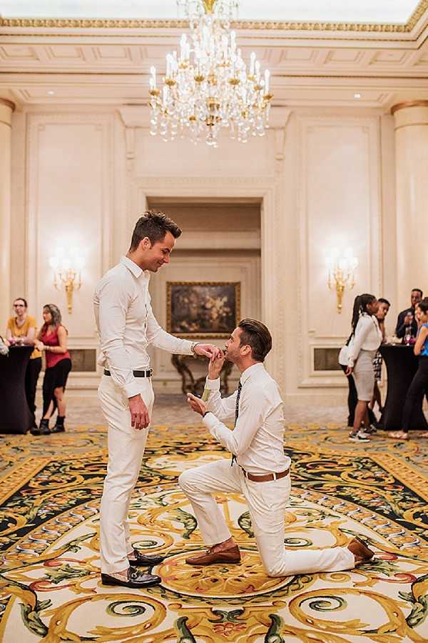 A marriage proposal is taking place inside a grand ballroom, where one man kneels on one knee presenting a ring to his partner who stands smiling above him. Both men are dressed in all-white outfits — white button-down shirts and white trousers — one wearing a dark tie with a brown belt, the other with a black belt. The setting is a formal, classically decorated ballroom with cream and gold molded walls, wall sconces, a large crystal chandelier overhead, and an ornate gold and black patterned carpet. Several guests are visible socializing in the background, unaware of or witnessing the moment from a distance. The shot is a full-length portrait taken from a medium distance, capturing both figures and the surrounding room context.