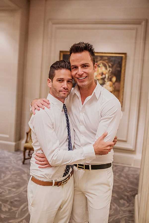 Two grooms pose together in a close embrace inside what appears to be a classic ballroom or hotel venue, with paneled cream walls and a framed oil painting visible in the background. Both men wear white dress shirts and cream/ivory trousers — one has a navy blue polka-dot tie and a tan leather belt, while the other wears no tie and a black belt. The setting has a formal, classic interior style with patterned carpeting and traditional molding. This is a mid-shot portrait with warm ambient indoor lighting.