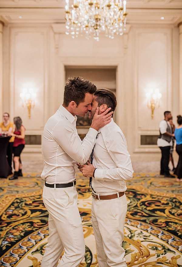 Two grooms share a kiss on the dance floor during what appears to be a wedding reception inside a formal ballroom. Both men are dressed in matching white dress shirts and white trousers — one with a black belt and the other with a tan leather belt — giving the overall look a coordinated, all-white style. The ballroom features warm wall sconces, a large crystal and gold chandelier overhead, ornate wall paneling, and a bold decorative carpet with gold and green floral motifs. Several guests are visible in the background, dressed in casual to semi-formal attire, mingling near the edges of the room. The image is a mid-range portrait shot with the couple in sharp focus and the background softly blurred.