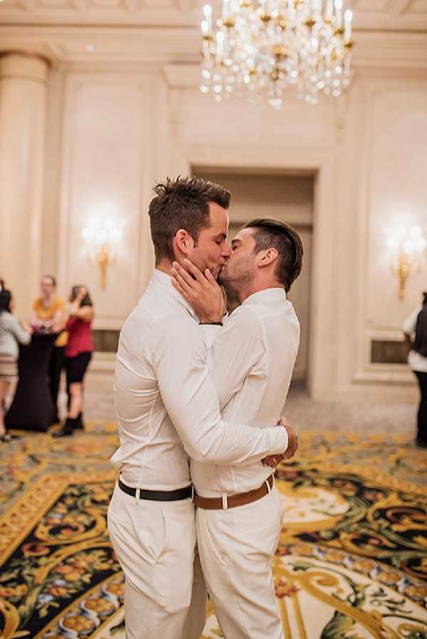 Two grooms share a kiss during what appears to be their first dance or a reception moment inside a formal ballroom. Both men are dressed in matching white dress shirts and white trousers — one with a black belt, the other with a tan leather belt — giving a coordinated, classic look. The interior space features ornate molded walls, gold wall sconces with warm lighting, a large crystal chandelier overhead, and a bold patterned carpet in black, gold, and green with a floral motif. Several guests are visible in the soft-focus background. The shot is a mid-range portrait taken at floor level, with warm ambient lighting creating a golden tone throughout the image.