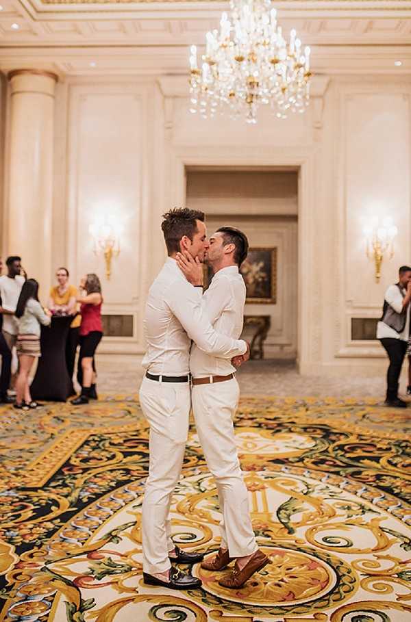 Two grooms share a kiss in the center of an ornate hotel ballroom or grand hall, both dressed in all-white outfits — white dress shirts and white trousers — with brown leather belts and tan loafers. The setting is a formal, classic interior featuring a large crystal chandelier overhead, gold wall sconces, decorative molded ceiling panels, and an elaborate patterned carpet in gold, black, and coral tones. Several guests in casual to smart-casual attire are visible in the background along the perimeter of the room. The shot is a full-length portrait taken from a low-to-mid angle, with the couple sharply in focus and the background slightly softened.