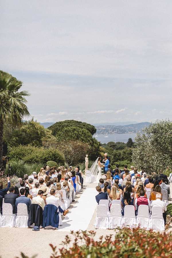 An outdoor wedding ceremony taking place on a wide terrace or garden path lined with white chairs on both sides, with approximately 80–100 guests seated facing the altar. The bride is visible in a white gown with a long cathedral veil, standing beside the groom who is wearing a bright cobalt blue suit, with an officiant nearby. The setting is a formal Mediterranean garden with palm trees, umbrella pines, and olive trees surrounding the space. A wide bay and coastline are visible in the background, suggesting a South of France location such as the Saint-Tropez peninsula. The composition is a wide elevated shot taken from behind the guests, capturing the full length of the ceremony aisle and the landscape beyond. Potential venue feature image.