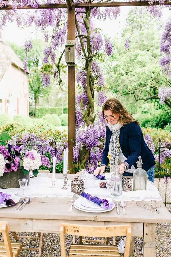 A wedding planner or florist is setting a reception table outdoors under a pergola draped with blooming purple wisteria. The rustic wooden table is dressed with a white linen runner and styled with white plates, silver cutlery, purple rolled napkins, silver candelabras with white taper candles, and glass siphon-style bottles. Floral centerpieces feature deep purple calla lilies, white hydrangeas, and purple lilacs arranged in dark vessels, tying into the purple-and-silver decor palette. The shot is a medium wide portrait capturing the woman leaning over the table mid-setup, with natural garden surroundings and a rustic building visible in the background.