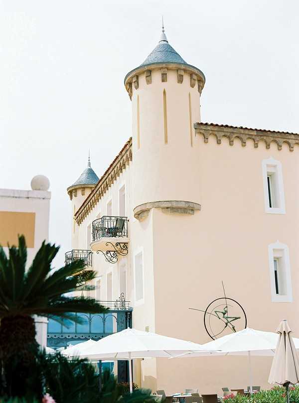 Wide shot of a chateau-style venue building featuring pale peach rendered walls, two conical-roofed turrets with slate-grey caps, decorative crenellations, and ornate wrought-iron balconies. In the foreground, white market umbrellas and outdoor seating are visible, suggesting a terrace or cocktail area, along with a sculptural metal sundial or armillary sphere. No people are visible in the frame. Potential venue feature image.