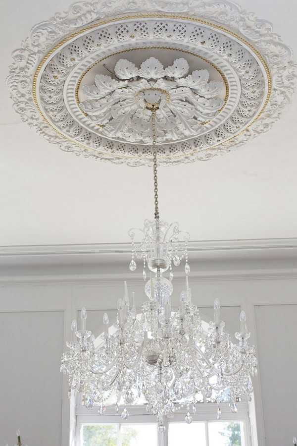 Detail shot looking up at an ornate white plaster ceiling medallion with gold accent trim, from which hangs a large multi-arm crystal chandelier with candle-style bulbs and hanging crystal drops. The room features white walls with classical crown molding, and a tall white-framed window is partially visible in the background. The interior architectural style is classic French chateau, with the all-white and gold palette creating a formal, light-filled setting. Potential venue feature image.