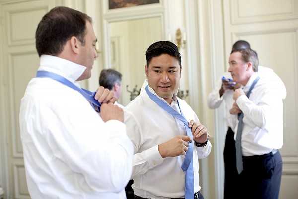 Getting-ready scene showing three men in white dress shirts adjusting their light blue ties before the ceremony. The setting appears to be an indoor room with white paneled walls and ornate mirrors, consistent with a chateau or formal venue interior. The men are at various stages of tying their neckties, with the central figure — likely the groom or a groomsman — focused on knotting his pale blue tie. Medium shot with a candid, documentary-style composition.