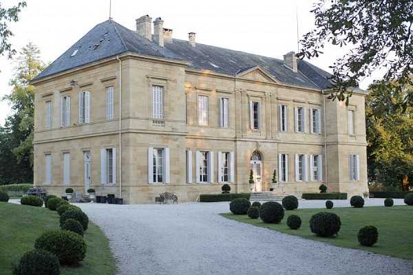 Wide exterior shot of a classic French château, a two-story limestone building with a slate mansard roof, white shuttered windows, and a central arched entrance door with interior lighting visible. The grounds feature a gravel driveway flanked by neatly clipped spherical boxwood topiaries and manicured lawn. No people are present in the image. Potential venue feature image.