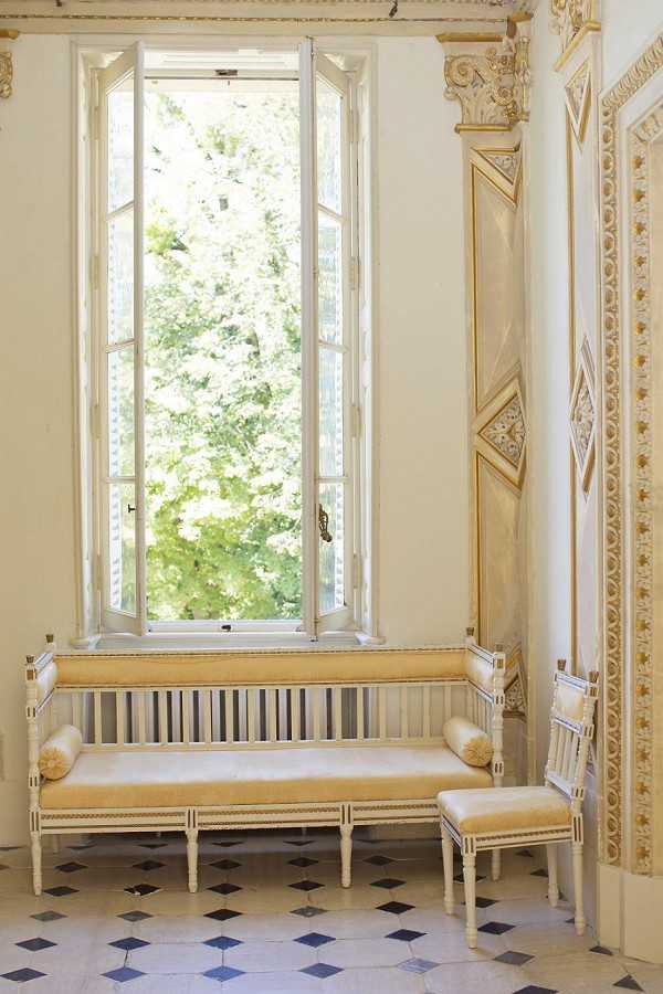Interior architectural detail shot of a formal French chateau or manor room, featuring a cream-painted wooden settee with gold velvet upholstery and matching bolster cushions positioned beneath a tall, open casement window. A small matching side chair is visible to the right. The room features ornate gold-accented plasterwork on the pilasters and wall panels, a black and white diamond-patterned marble floor, and white walls with classical carved column capitals. No people are present in the image. The composition is a medium wide shot emphasizing the period interior decor with a classic French style palette of cream, gold, and blue-grey. Potential venue feature image.